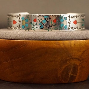 May include: A silver cuff bracelet with a floral design. The bracelet is decorated with turquoise, red, and white stones.