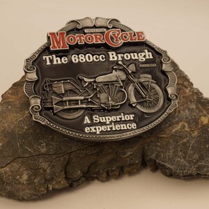 May include: A motorcycle belt buckle with the text "MOTOR CYCLE" in red and a motorcycle design. The buckle is dark gray with silver accents and the text "The 680cc Brough" and "A Superior experience".