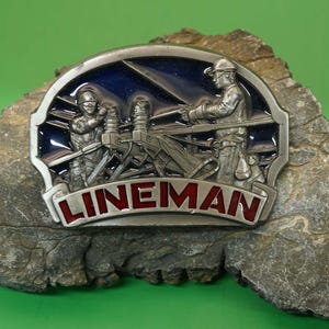 Op de afbeelding: Een zilveren gesp met een blauw en rood emaille ontwerp dat twee monteurs toont die aan een stroomlijn werken. De gesp heeft het woord "LINEMAN" in rode letters.