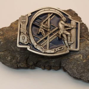 Op de afbeelding: Zilverkleurige metalen riemgesp met een blauwe emaille achtergrond, met twee figuren die aan een constructie werken. De gesp heeft een rechthoekige vorm met een gedetailleerd ontwerp, inclusief een telefoonpaal en een ketting.