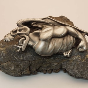 May include: A silver-colored gargoyle sculpture with detailed wings and a skeletal head, resting on a rough, gray stone. The sculpture has a muscular body and is likely a decorative item.