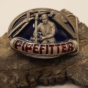 Op de afbeelding: Een zilveren gesp met een blauw en rood emaille ontwerp. De gesp toont een loodgieter die aan een pijp werkt met gereedschap. De tekst "PIPEFITTER" staat in rood op de gesp.