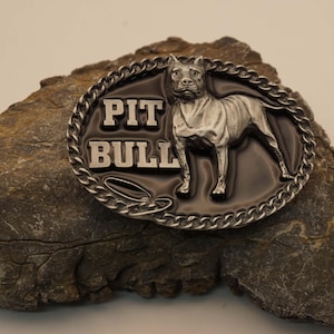 May include: An oval-shaped metal belt buckle featuring a raised silver-toned Pit Bull dog figure and the text PIT BULL in block letters against a smooth black background, all framed by a decorative silver-toned chain-link border design.