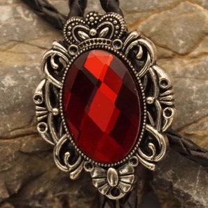 Può includere: Un bolo tie argentato con una pietra preziosa rossa incastonata in una cornice decorativa. La cornice presenta un design a corona e dettagli intricati.