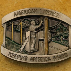 Op de afbeelding: Zilveren gesp met een groene, bruine en gouden afbeelding van een monteur die aan een elektriciteitsmast werkt. De gesp heeft de tekst "American Lineman Keeping America Wired".
