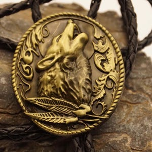 Può includere: Un bolo tie dorato con un designo dettagliato di testa di lupo con intricati accenti floreali. Il lupo sta ululando a bocca aperta e con la testa inclinata all'indietro. Il bolo tie è fatto di metallo e ha un cordino in pelle nera.