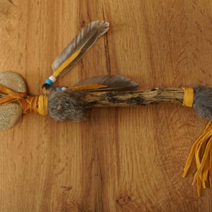 Könnte beinhalten: Eine Friedenspfeife im indianischen Stil mit einem Steinkopf, einem Holzschaft und einem grauen Fellquaste. Der Schaft ist mit gelbem Leder umwickelt und mit Federn und Perlen verziert.