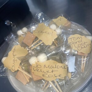 Puede incluir: Primer plano de una colección de pequeñas muestras de perfume en bolsas de plástico transparentes. Cada muestra tiene un pequeño palo de madera y una bola de algodón blanca. Las bolsas están etiquetadas con nombres escritos a mano, incluyendo "Creed", "Tom Ford Lost Cherry", "Jo Malone Velvet Rose" y "Olympea".