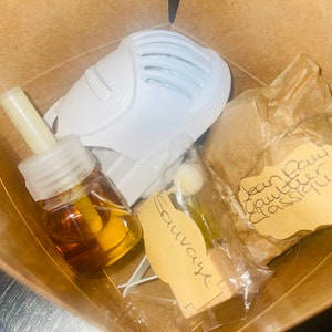 Puede incluir: Una caja de cartón marrón con un difusor blanco, una pequeña botella de líquido ámbar, un pequeño bloque de madera con la palabra "Sauvage" escrita en él y una bolsa de papel marrón con el texto "Jean Paul Gaultier Classique" escrito en ella.