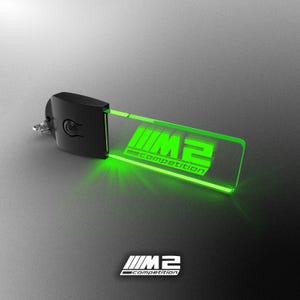 May include: A black keyring with a rectangular, transparent, illuminated section. The text "M2 COMPETITION" is displayed in green. The keyring is on a gradient grey background.