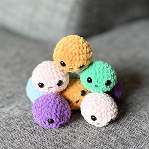 May include: A collection of handmade, colorful crochet balls with black safety eyes and stitched smiles. The balls are in shades of yellow, pink, green, purple, and blue. These plush toys are soft and cuddly.