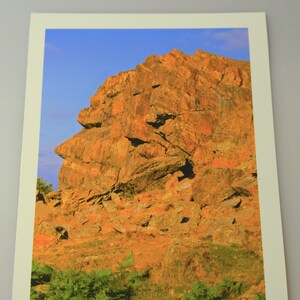 Pode incluir: Uma impressão de paisagem com uma grande formação rochosa castanho-avermelhada sob um céu azul claro. A rocha tem uma superfície texturizada com sombras e fendas. A folhagem verde é visível na base da formação rochosa.