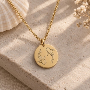 May include: A gold-toned necklace featuring a circular pendant engraved with two whale sharks. The pendant hangs from a delicate matching gold-toned chain, showcasing a detailed line art design of the sharks swimming in a circular pattern.