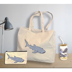May include: A set of three whale shark themed items: a tote bag, a makeup bag, and a water bottle. The tote bag is made of natural canvas and features a blue whale shark with white spots. The makeup bag is a smaller version of the tote bag with the same design. The water bottle is white with a blue whale shark design and a bamboo lid.