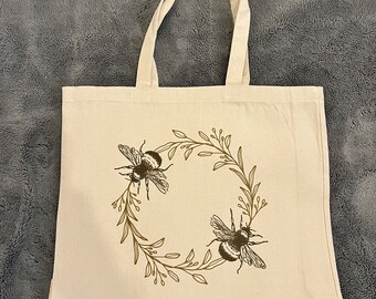 May include: A beige canvas tote bag with brown floral and bee design. The bees are in a circle with leaves and branches.