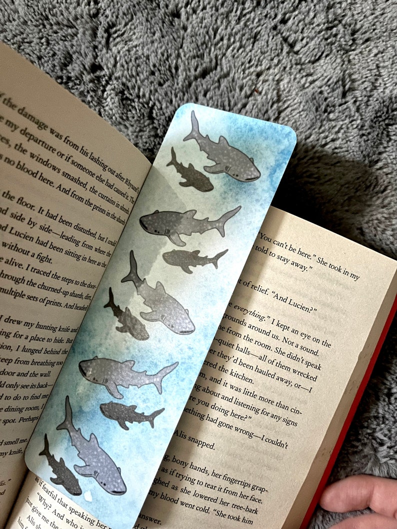 May include: A blue and white bookmark with a watercolour design of several grey whale sharks swimming in a blue ocean. The bookmark has text on the right side that reads "You can't be here. She took in my told to stay away."