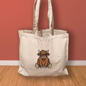 May include: A white canvas tote bag featuring a brown cartoon Highland cow sitting on the front. The cow has a black nose and hooves.