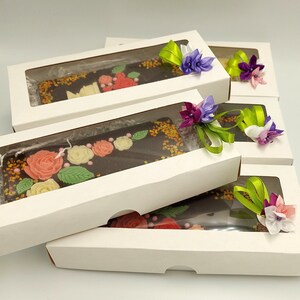 Op de afbeelding: Verschillende chocoladerepen in witte dozen met doorzichtige ramen. De donkere chocoladerepen zijn versierd met kleurrijke bloemmotieven en sprinkles. Elke doos is versierd met een lint en een bloemstuk in paars, groen en wit.