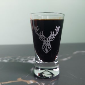 May include: A clear shot glass filled with a dark brown liquid. The glass features a silver geometric deer head design. The glass sits on a gray and white marble surface.