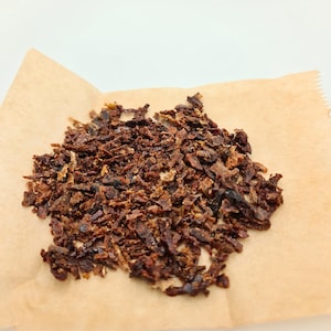 Può includere: Un mucchio di pezzi scuri e marroni sminuzzati, possibilmente cibo, poggia su un quadrato di carta marrone chiaro. I pezzi hanno una consistenza ruvida e diverse sfumature di marrone, suggerendo che potrebbero essere essiccati o cotti.