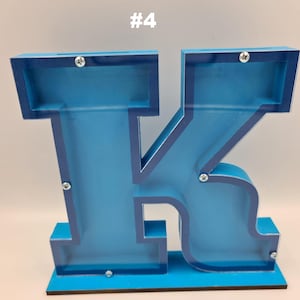 Può includere: Un salvadanaio a forma di lettera K blu con un pannello frontale trasparente e viti argentate visibili. La lettera K è montata su una base blu piatta. Il testo #4 è visibile sopra la lettera. Il design presenta una fessura sulla parte superiore.