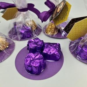 Può includere: Diverse piccole caramelle avvolte in carta viola sono disposte su cerchi viola. Alcune sono in sacchetti di cellophane trasparente legati con nastro viola e una piccola etichetta. Alcune caramelle sono avvolte in carta dorata.