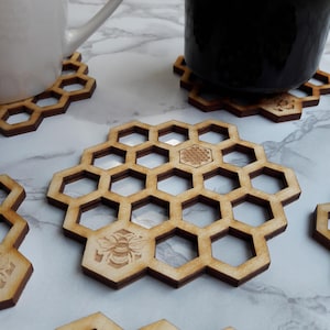 Könnte beinhalten: Holzuntersetzer in Wabenform mit gelaserten Bienen- und Blumendesigns. Die Untersetzer sind hellbraun und haben ein sechseckiges Muster. Zwei Tassen, eine weiße und eine schwarze, stehen auf den Untersetzern. Die Untersetzer haben einen Durchmesser von etwa 10 cm.