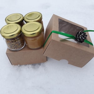 Può includere: Quattro piccoli vasetti di vetro con coperchi dorati, pieni di miele e polline d'api, sono su una scatola di cartone marrone. Una seconda scatola con finestra e nastro verde è accanto ai vasetti. Una pigna decora la scatola.