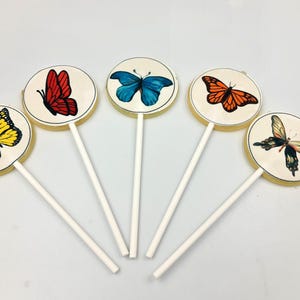 Könnte beinhalten: Fünf runde Lollis mit Schmetterlingsmotiven. Jeder Lolli zeigt einen anderen Schmetterling in leuchtenden Farben: Gelb, Rot, Blau, Orange und Schwarz. Die Schmetterlinge sind auf einem cremefarbenen Hintergrund mit einem weißen Stiel zentriert.
