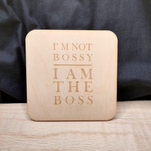 Könnte beinhalten: Quadratischer Holzuntersetzer mit dem eingravierten Text "I'M NOT BOSSY I AM THE BOSS". Der Untersetzer ist aus hellem Holz gefertigt und liegt auf einer Holzoberfläche. Der Hintergrund ist ein dunkler Stoff.