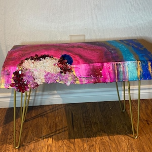 May include: A handmade coffee table with an abstract design. The tabletop features pink, blue, and gold colours, decorated with crystal embellishments. Supported by gold hairpin legs, this artistic piece adds a touch of elegance. The table measures approximately 100 cm in length.