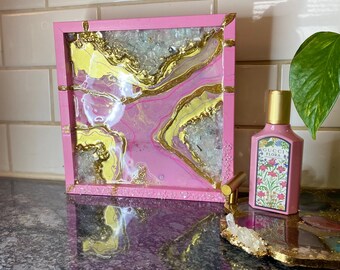 Könnte beinhalten: Ein rosa Harztablett mit goldenen und gelben Akzenten. Das Tablett hat eine rechteckige Form und ist mit einem fließenden Design verziert. Eine rosa Gucci Flora Parfümflasche und ein dekoratives Tablett sind ebenfalls sichtbar.