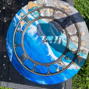 May include: Circular wall clock with a blue and white wave design. The clock face has Roman numerals and is decorated with seashells. The clock has a dark metal frame and black hands. A unique coastal home decor item, approximately 30 cm in diameter.