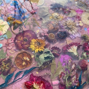 May include: Close-up of a resin art piece with dried flowers and citrus slices embedded in a pink background. The flowers are in shades of purple, yellow, and red. Gold leaf flakes add a touch of sparkle to the artwork.