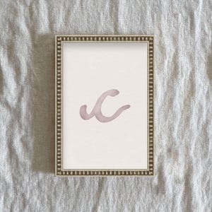 May include: A framed print with a watercolor design of a pink abstract shape resembling a letter C. The frame is silver with a beaded border.