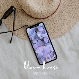 以下が含まれることがあります： スマートフォンには紫色の紫陽花のクローズアップが表示されています。 電話は、麦わら帽子と黒いサングラスの隣の白い表面に置かれています。 画像の下部には「bloom house PRINT SHOP」というテキストがあります。