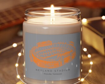 May include: A lit candle in a clear glass jar with a gray label featuring an orange illustration of Neyland Stadium. The text "NEYLAND STADIUM Knoxville, Tennessee" is below the stadium image. The candle sits on a wooden coaster.