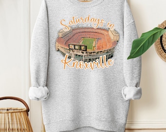 Può includere: Una felpa grigio chiaro con la scritta "Saturdays in Knoxville" in arancione. La grafica raffigura uno stadio con toni arancioni e marroni. La felpa è appesa a una gruccia di legno.