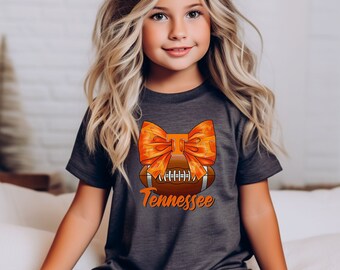 Puede incluir: Una camiseta gris oscuro con un diseño de fútbol americano, un lazo naranja y la palabra "Tennessee" en escritura naranja. La camiseta la lleva una niña con el pelo largo y rubio.