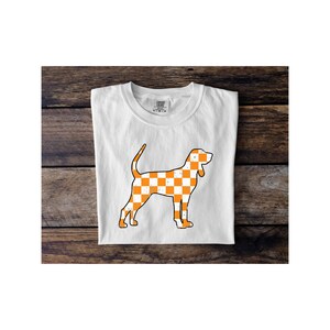 May include: White t-shirt featuring a stylized orange and white checkered dog graphic. The dog illustration has a distressed, vintage look. The shirt is folded neatly on a wooden surface.