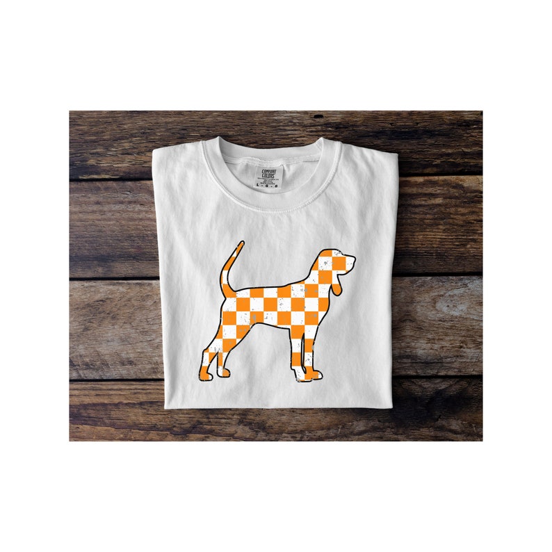 May include: White t-shirt featuring a stylized orange and white checkered dog graphic. The dog illustration has a distressed, vintage look. The shirt is folded neatly on a wooden surface.