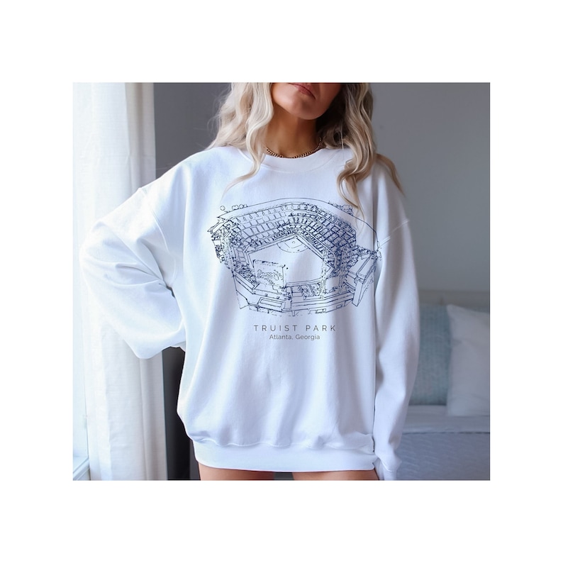 K&ouml;nnte beinhalten: Wei&szlig;er Pullover mit einer blauen Umrandung des Truist Park, einem Baseballstadion in Atlanta, Georgia. Der Text "Truist Park" und "Atlanta, Georgia" ist unterhalb der Stadionumrandung aufgedruckt.