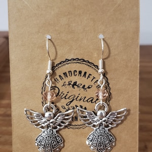 May include: Silver angel dangle earrings with a peach-colored bead on each earring. The earrings are on a brown card with the text "HANDCRAFTED ORIGINAL".