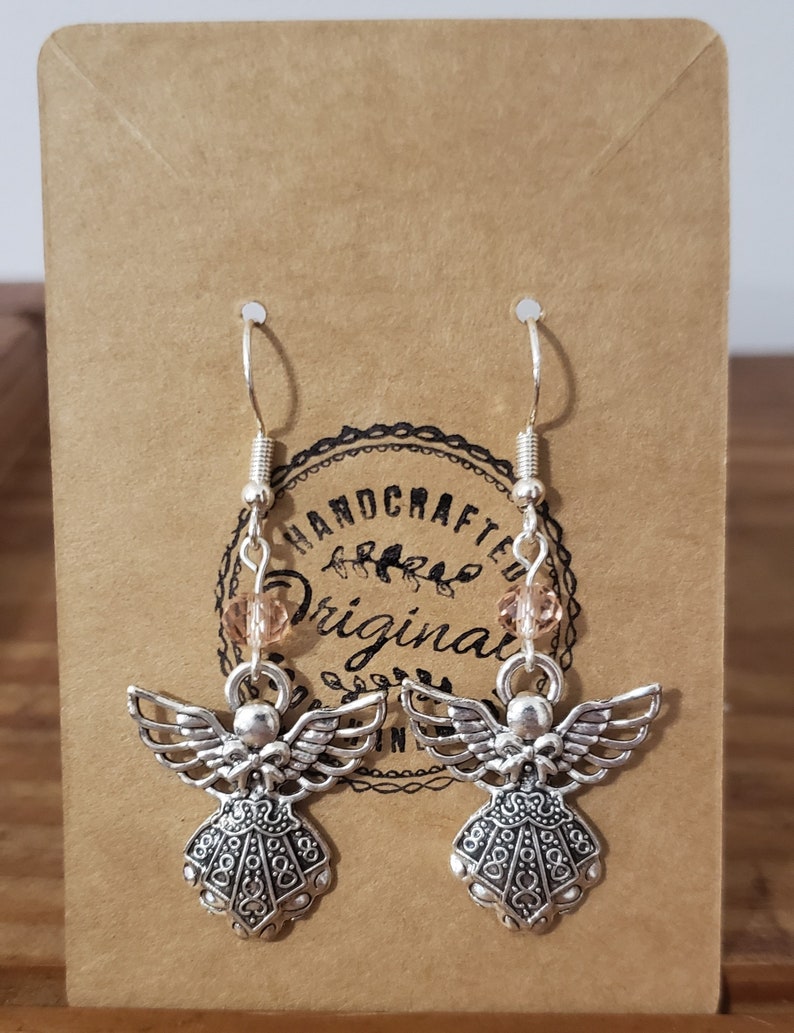 May include: Silver angel dangle earrings with a peach-colored bead on each earring. The earrings are on a brown card with the text "HANDCRAFTED ORIGINAL".