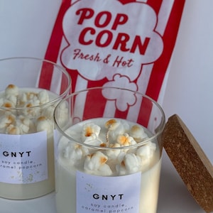 Op de afbeelding: Twee glazen kaarsen gevuld met witte was en gegarneerd met popcorn. Elke kaars heeft een label met de tekst "GNYT soy candle, caramel popcorn". Op de achtergrond een rood-witte popcornzak.