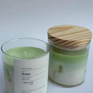 May include: Two green and white soy wax candles in glass jars with wooden lids. The label on one jar reads "Gnyt Candles", "Order", "June 6, 2024", "4:58:12 PM", "1x Matcha Latte", "Large", "coffee is coffee, but if you make it well, it can be art.", "Small soy coconut apricot candle hand poured in Dublin, OH."
