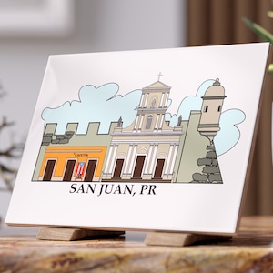 May include: A white ceramic tile featuring a colorful illustration of San Juan, PR. The artwork depicts buildings, a church, and a fort against a light blue sky. The tile is displayed on a wooden stand, perfect for home decor.