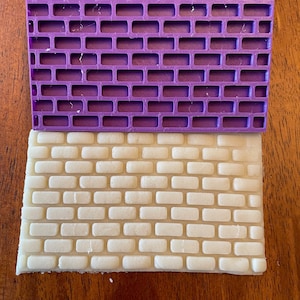 May include: A purple plastic brick mold with a white brick pattern pressed into it. The mold is on top of a sheet of white fondant.