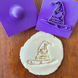 May include: Purple plastic cookie cutters in the shape of a witch's hat. The cutters are on a wooden surface with a white play dough imprint of the hat.