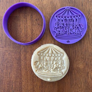 May include: A purple circular cookie cutter and a matching purple stamp featuring a detailed carousel design with horses. Below the tools, a round cookie shows the embossed carousel image, complete with a small star at the bottom edge.